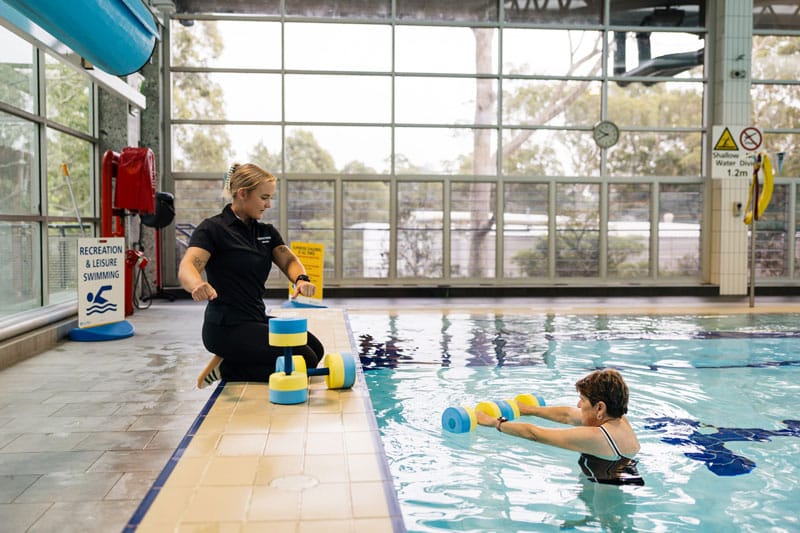 Physiotherapist guiding an older female client through a hydrotherapy session using foam dumbbells in an indoor pool.
