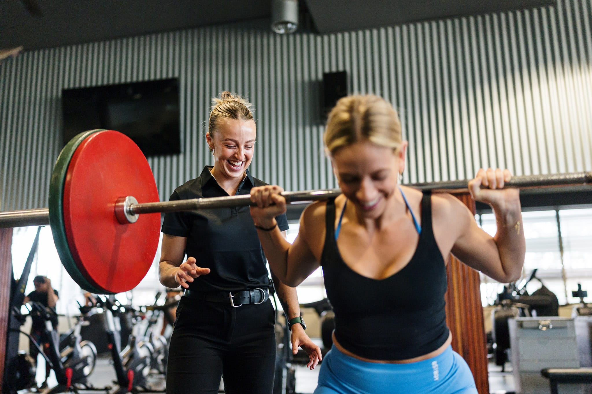 Physiotherapist coaching a female client during a squat with a barbell in a gym setting, both smiling and engaged.